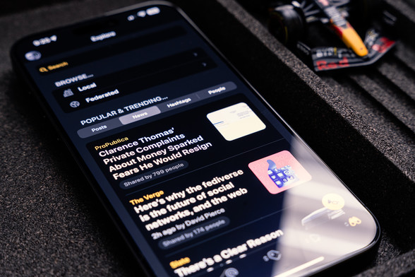 Photo of an iPhone with the new Explore tab shown on screen. Shows Search, Browse (Local/Federated), and Popular & Trending Posts, News, Hashtags, and People.